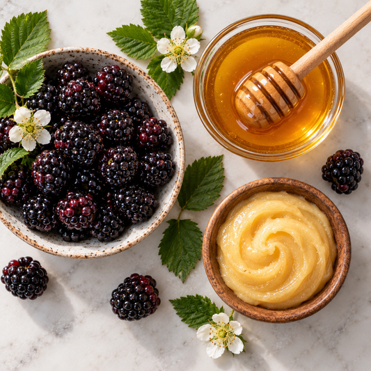 A styled flat-lay of a blackberry and honey product, featuring a bowl of fresh blackberries with white flowers, a bowl of smooth golden honey, and a bowl of blackberry jam on a light marble surface.